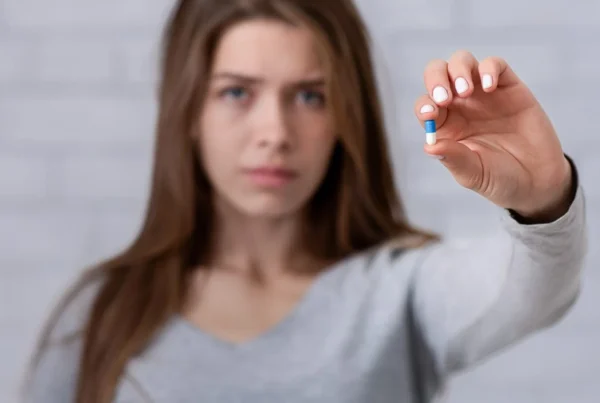 Depressed young lady showing antidepressant or sedative tablet against white brick wall, selective focus.