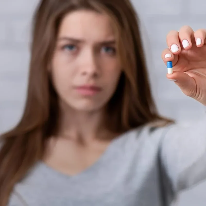 Depressed young lady showing antidepressant or sedative tablet against white brick wall, selective focus.
