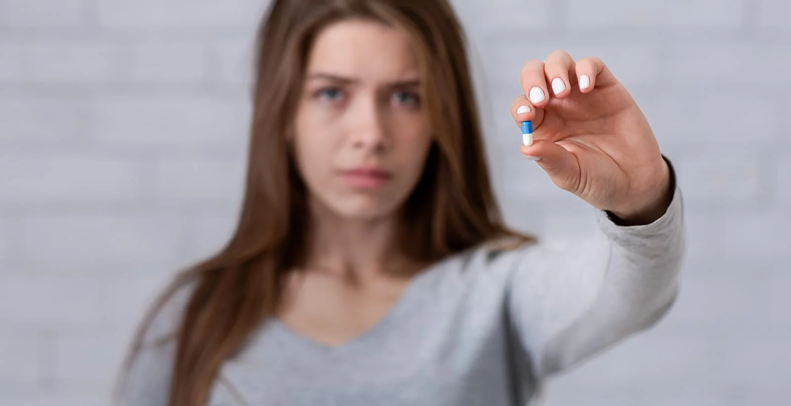 Depressed young lady showing antidepressant or sedative tablet against white brick wall, selective focus.